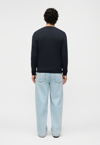 Άνδρας με σγουρά μαλλιά που φορά μονόχρωμο μαύρο μακρυμάνικο πουκάμισο και φαρδιά ανοιχτό μπλε τζιν, στέκεται γυρισμένος με την πλάτη προς λευκό φόντο.