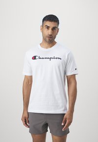 Vit bomullströja med en svart och röd "Champion"-logga tvärs över bröstet, korta ärmar, rund halsringning och minimalistisk design.