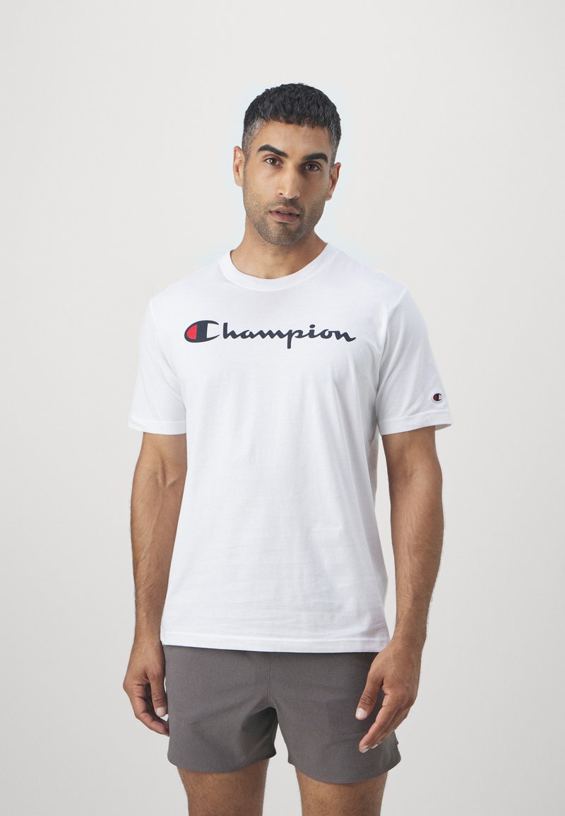 Vit bomullströja med en svart och röd "Champion"-logga tvärs över bröstet, korta ärmar, rund halsringning och minimalistisk design.