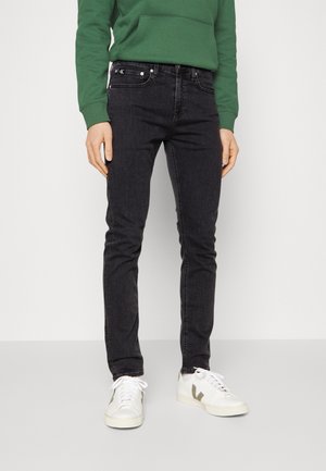 Persoon die nauwsluitende zwarte jeans, witte sneakers met bruine details en een groene trui draagt, staand tegen een effen lichte achtergrond.