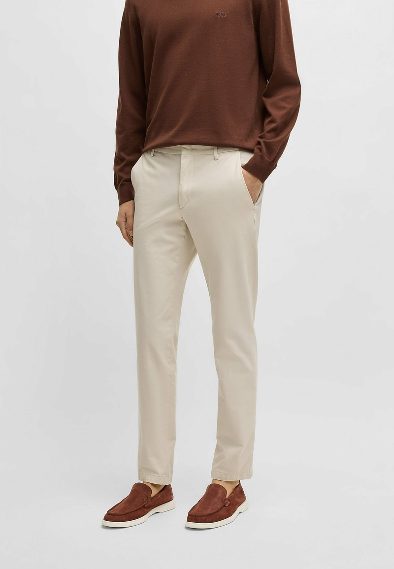 Beige tailored pants with side pockets, paired with a brown long-sleeve shirt. The shoes are brown suede loafers with a white sole.