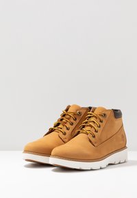 Botas de tobillo de ante marrón con cordones amarillos, suela blanca y detalles en marrón. Presentan una puntera redondeada y una superficie texturizada.