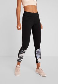 Kobieta w czarnych legginsach z kwiecistymi wstawkami na łydkach i beżowych butach sportowych, stojąca na jednolitym tle.