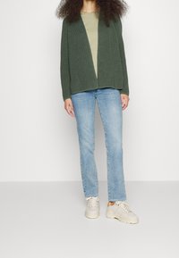 Olive green knit cardigan with open front, textured design. Paired with light blue straight-leg jeans and beige chunky sneakers.