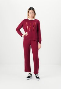 Woman stands against white background wearing burgundy GAP sweatshirt and matching pants with black and white sneakers.
