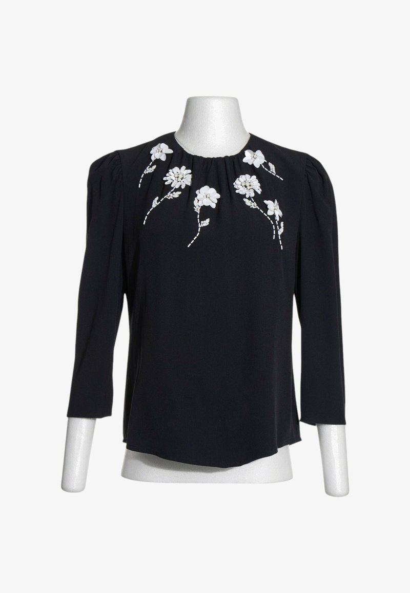 Blouse noire à manches longues avec épaules bouffantes ornée d'une broderie florale blanche autour de l'encolure sur un mannequin blanc.