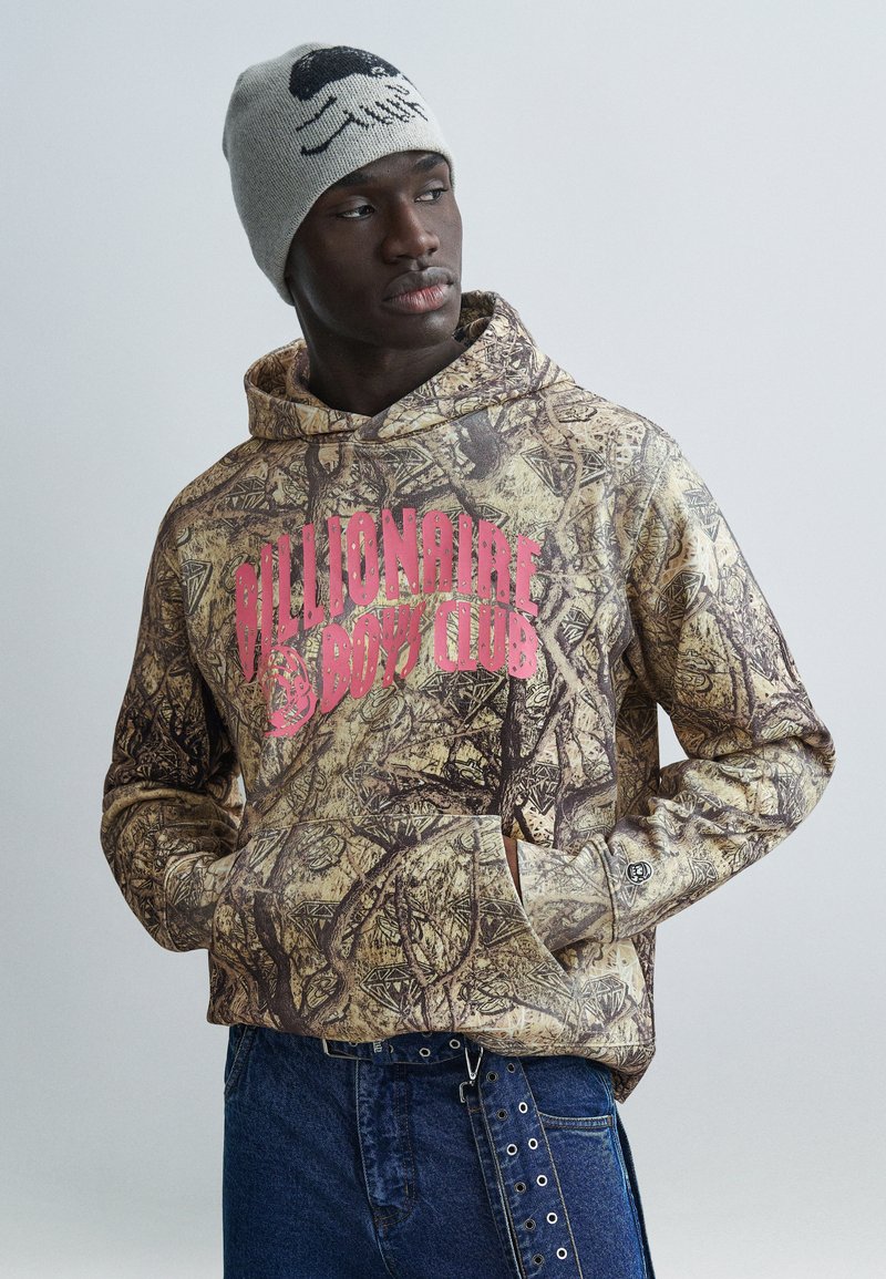 Φούτερ με κουκούλα σε καμουφλάζ κα φέ και μπεζ χρώματα, με έντονο ροζ λογότυπο "Billionaire Boys Club". Φοράται με σκούρο τζιν και γκρι σκουφάκι.