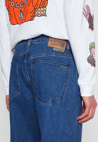 Jeans de denim azul con un corte relajado, que presentan un parche de cuero marrón y una pequeña etiqueta con el logo. La tela muestra un tejido texturizado clásico.