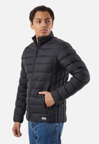 Jeune homme portant une veste matelassée noire avec des poches zippées et un jean bleu, regardant sur le côté sur un fond blanc.