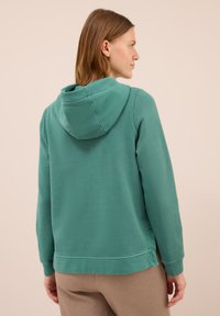 Mujer con cabello castaño claro, vistiendo una sudadera color teal y pantalones beige, mostrada de espaldas y ligeramente girada hacia un lado.