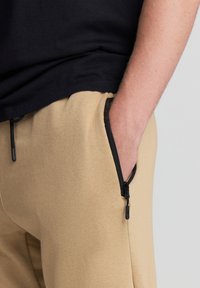 Pantaloni della tuta beige con tasche laterali con zip, dotati di una vita regolabile con cordino, texture morbida e dettagli in zip nera a contrasto.