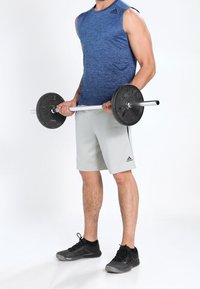 Man in blue sleeveless shirt and gray shorts holding a barbell with weights, standing on a white floor with black athletic shoes.