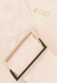 Prodotto in pelle rosa chiaro con superficie testurizzata, dotato di una fibbia in metallo dorato e di una tracolla in nastro con lettering impressi.