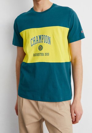 Man in een teal en gele Champion T-shirt met de tekst "ROCHESTER 1919", gecombineerd met beige broek, staand tegen een witte achtergrond.