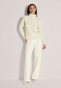 Vrouw in een off-white geribbelde coltrui met wijde crèmekleurige broeken en witte puntige hakken, geposeerd tegen een effen achtergrond.