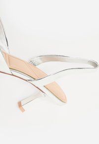 Stříbrné sandály na vysokém podpatku s elegantním designem, vybavené tenkými pásky a texturovanou podrážkou, kontrastující s lehce béžovou stélkou.