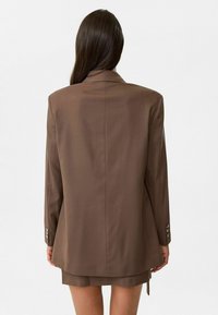 Bruine op maat gemaakte blazer met een gestructureerd ontwerp, voorzien van een enkele rugsplit en zilveren knoopaccenten op de mouwen. Gladde stoftextuur.