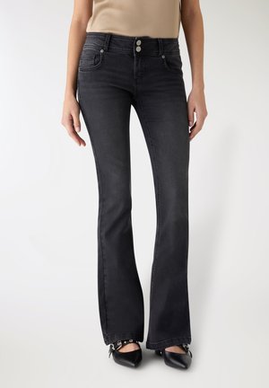 Donna che indossa jeans neri a vita alta svasati con chiusura a doppio bottone e scarpe nere con punta appuntita e fibbie, in piedi contro uno sfondo bianco.