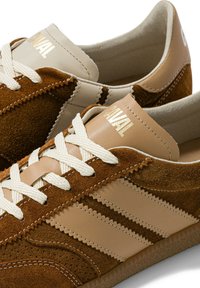 Baskets en daim marron et cuir beige avec lacets blancs et motif rayé sur le côté, montrant des détails en gros plan des coutures et de la texture.