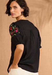 Fekete felső puffos ujjal, színes virágos hímzéssel a vállakon, texturált anyaggal, amelyen finom minták láthatók, és laza fazonnal.