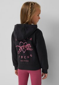 Zwarte hoodie met een roze graphic waarop een leeuw, een hart en de tekst "CIRCUS" op de achterkant zijn afgebeeld. Gepaarde met roze geribde leggings.