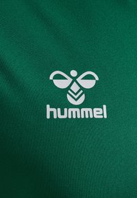 Groene textiel met een opvallend wit logo van een gestileerde bij en de tekst "hummel" eronder. Gladde textuur en een gelijkmatige stofweving.