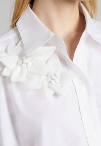Camisa de algodón blanca con cuello spread, que presenta dos grandes apliques florales blancos en el lado izquierdo. Detalle de botones en la parte delantera.