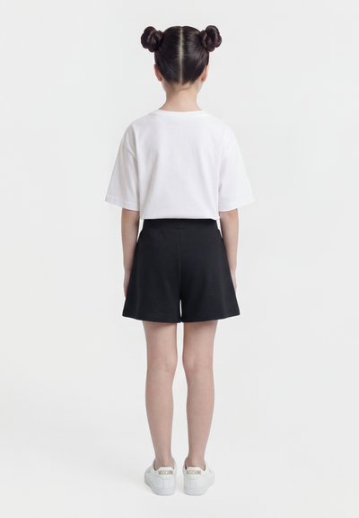 Enfant debout de dos, portant un t-shirt blanc, un short noir, des baskets blanches, les cheveux coiffés en deux chignons.