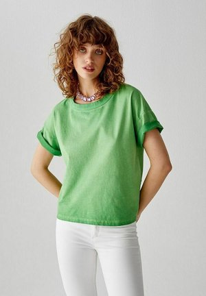 T-shirt verde in cotone con maniche arrotolate, scollatura rotonda, texture morbida. Indossato con jeans bianchi attillati, dettagli di cuciture minimi visibili.