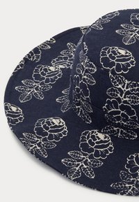 Marineblå hatt laget av strukturert stoff, med et blomsterhvitt mønster. Bred skjerm med rund kroneform, som gir solbeskyttelse.