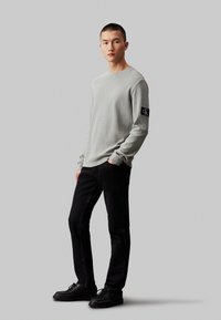 Camisa de manga larga gris texturizada con parche de logo en la manga izquierda, combinada con jeans ajustados negros y zapatos negros con cordones.