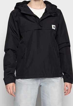 Persoon draagt een zwarte windjack met capuchon, ritszakken en een klein vierkant logo op de borst, gecombineerd met lichtgrijze jeans.