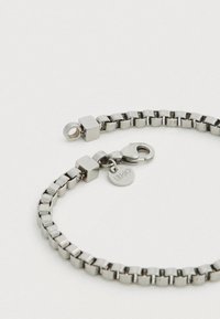 Brățară din metal argintiu cu zale pătrate, având o închizătoare tip homar și o etichetă rotundă. Textură netedă cu un finisaj strălucitor.