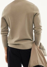 Béžový pletený svetr s dlouhými rukávy, ohrnutými manžetami a kulatým výstřihem, spolu s černými kalhotami, pod kterým je světle hnědá bunda.