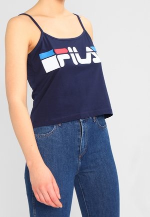 Vrouw die een marineblauwe Fila spaghetti-strap crop top en hoog getailleerde blauwe spijkerbroek draagt, staand tegen een effen achtergrond.