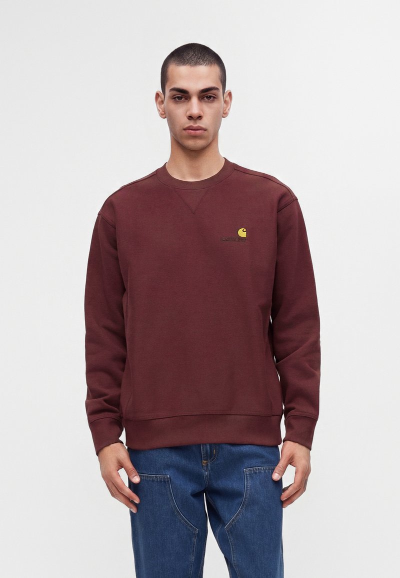 Jeune homme aux cheveux courts portant un sweat-shirt bordeaux avec un petit logo jaune et un jean bleu, debout devant un fond clair uni.