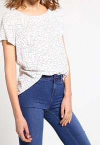 Haut blanc à manches courtes avec un motif de lignes noires dispersées, associé à un jean bleu taille haute. Coupe décontractée, texture lisse.