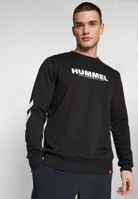 Svart hettegenser med lange ermer, med en hvit "HUMMEL"-logo på brystet og kontrasterende hvite detaljer på ermerne. Teksturert stoff.