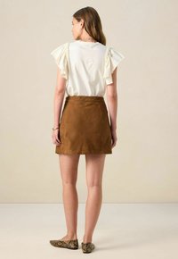 Brauner Wildleder-Minirock kombiniert mit einem cremefarbenen Oberteil mit gerüschten Ärmeln. Das Modell trägt gemusterte Ballerinas und wird von hinten betrachtet.