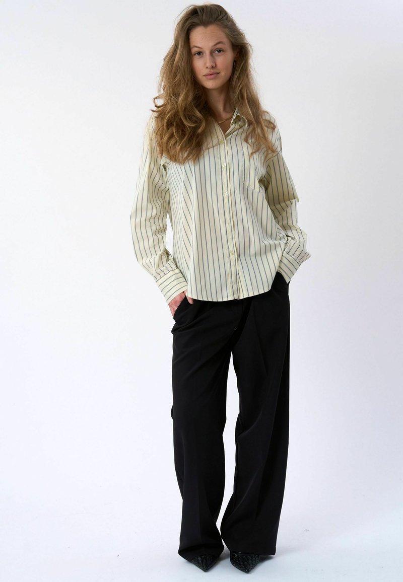 Jeune femme aux longs cheveux ondulés portant une chemise boutonnée rayée jaune clair, un pantalon noir à jambes larges et des chaussures noires pointues, debout de manière décontractée.