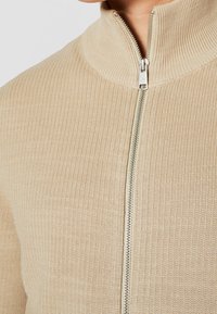Beżowy prążkowany sweter zapinany na zamek z wysokim kołnierzem i srebrnym detalem zamka, wykonany z teksturowanej, miękkiej tkaniny.