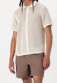 Pull polo à manches courtes en maille écru, doté d'une texture à tissage ouvert, d'une patte de boutonnage à l'avant et d'un col classique. Assorti avec un short marron.