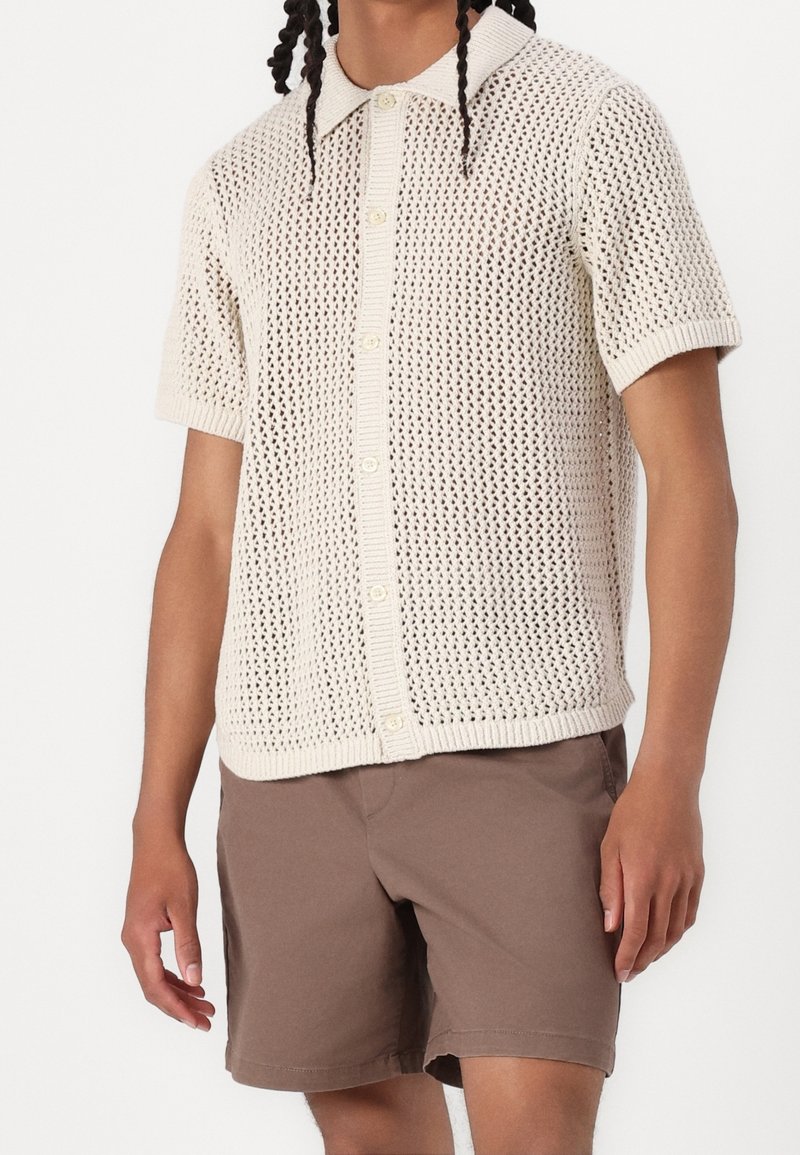 Pull polo à manches courtes en maille écru, doté d'une texture à tissage ouvert, d'une patte de boutonnage à l'avant et d'un col classique. Assorti avec un short marron.