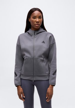 Žena v šedé mikině na zip s předními kapsami a malým černým logem Adidas, doplněná tmavými legínami, stojí před jednoduchým pozadím.