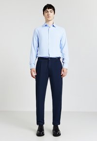 Junger Mann steht in einem hellblauen Hemd, blauen Hosen mit Knöpfen am Bund und schwarzen Schnürschuhen vor einem weißen Hintergrund.