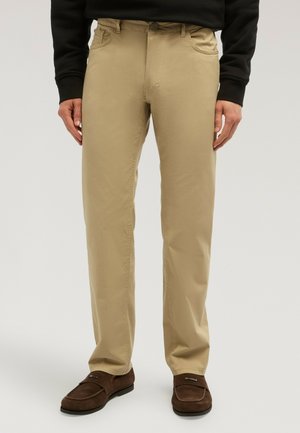 Mann trägt beige gerade geschnittene Hosen, dunkelbraune Mokassins und ein schwarzes Langarmoberteil, steht vor einem schlichten Hintergrund.