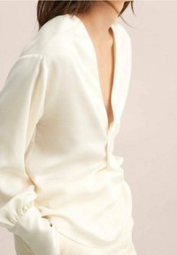 Cremfarbene Bluse mit tiefem V-Ausschnitt, langen Ärmeln mit gerafften Manschetten und einem glatten, strukturierten Stoff.