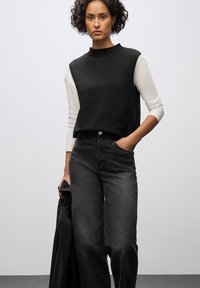 Top nero senza maniche sopra una camicia bianca a maniche lunghe, abbinato a jeans neri larghi. Il modello tiene una giacca nera. Stile semplice e moderno.