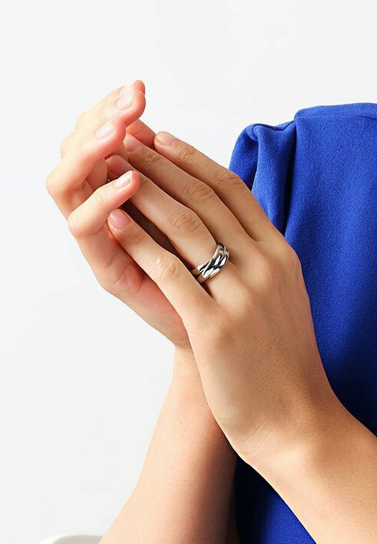 Ein geflochtener Ring aus Sterlingsilber, der an Fingern mit glatter Hauttextur getragen wird, die Hände sind vor einem blauen Stoffhintergrund gefaltet.