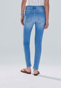 Világoskék szűk szabású farmernadrág halványított dizájnnal, két hátsó zsebbel és szabványos derékpánttal. Barna lapos szandállal viselve.